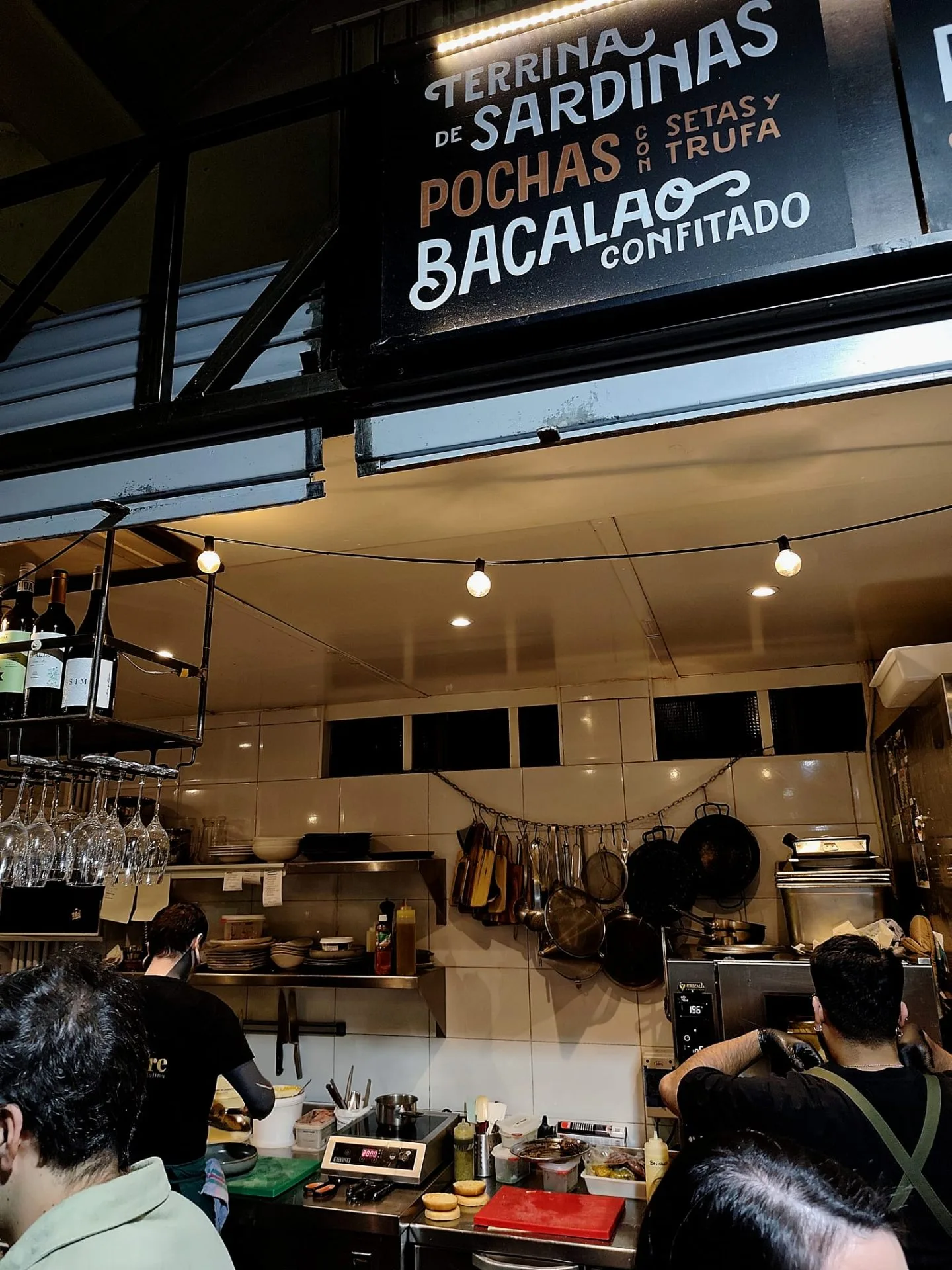 Cartel de terrina de sardinas, pochas con setas y trufa, y bacalao confitado sobre la cocina abierta del puesto de Silvestre en el Mercado de Tirso de Molina.